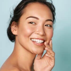 Asian woman with hair pinned back smiles while touching her cheek and admires glowing, youthful skin.